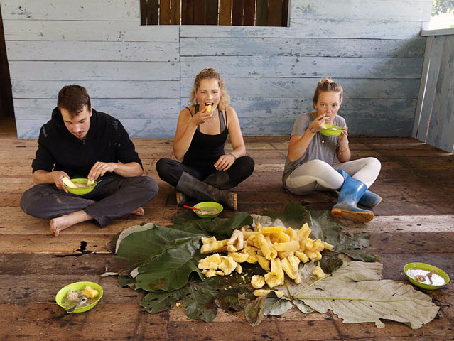 Drei Freiwillige sitzen im Schneidersitz auf dem Boden und essen Kochbananen und Suppe